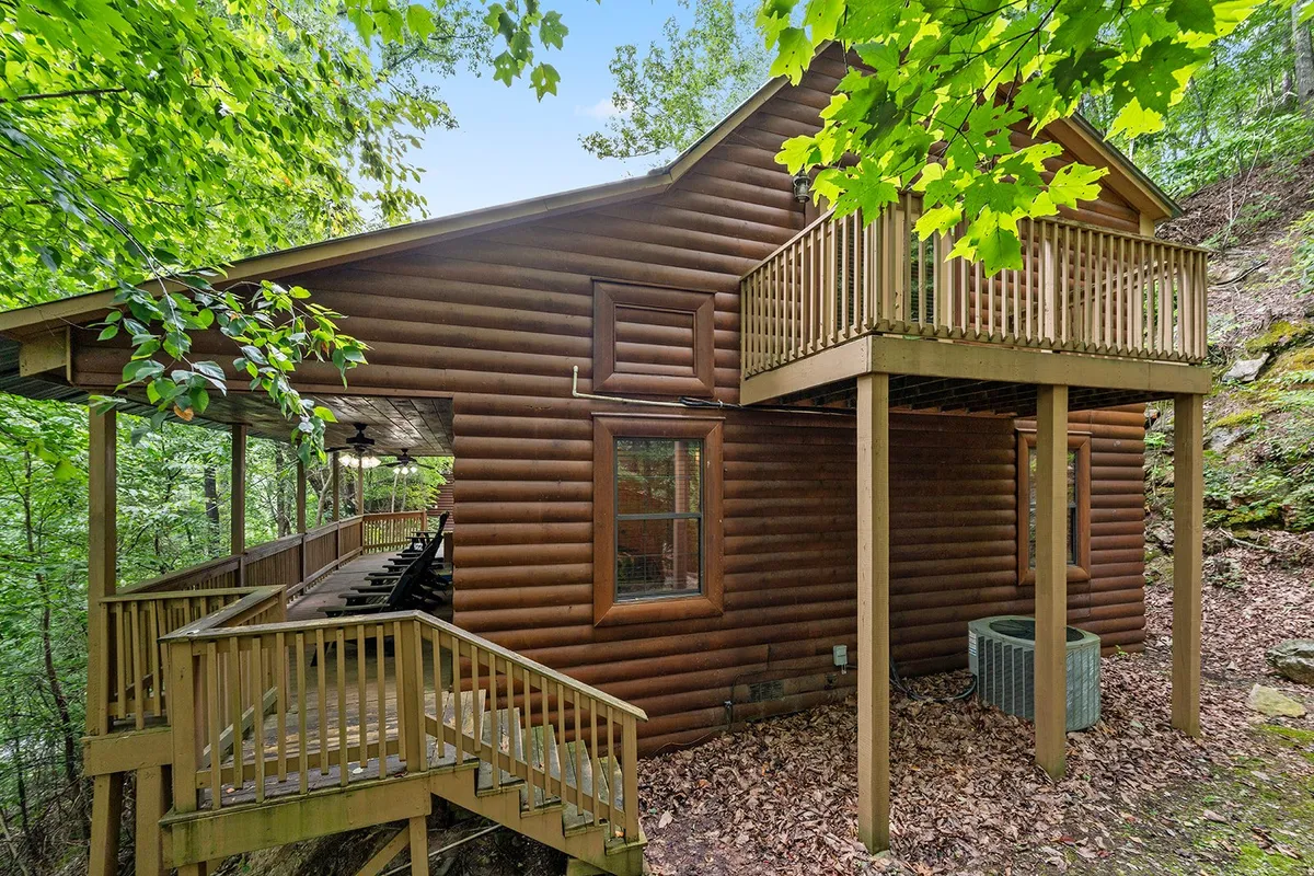 A log cabin with a wraparound deck and upper balcony set amidst lush foliage. The exterior emphasizes the spacious decks perfect for outdoor gatherings or relaxing, with ample space to enjoy mountain views and fresh air.