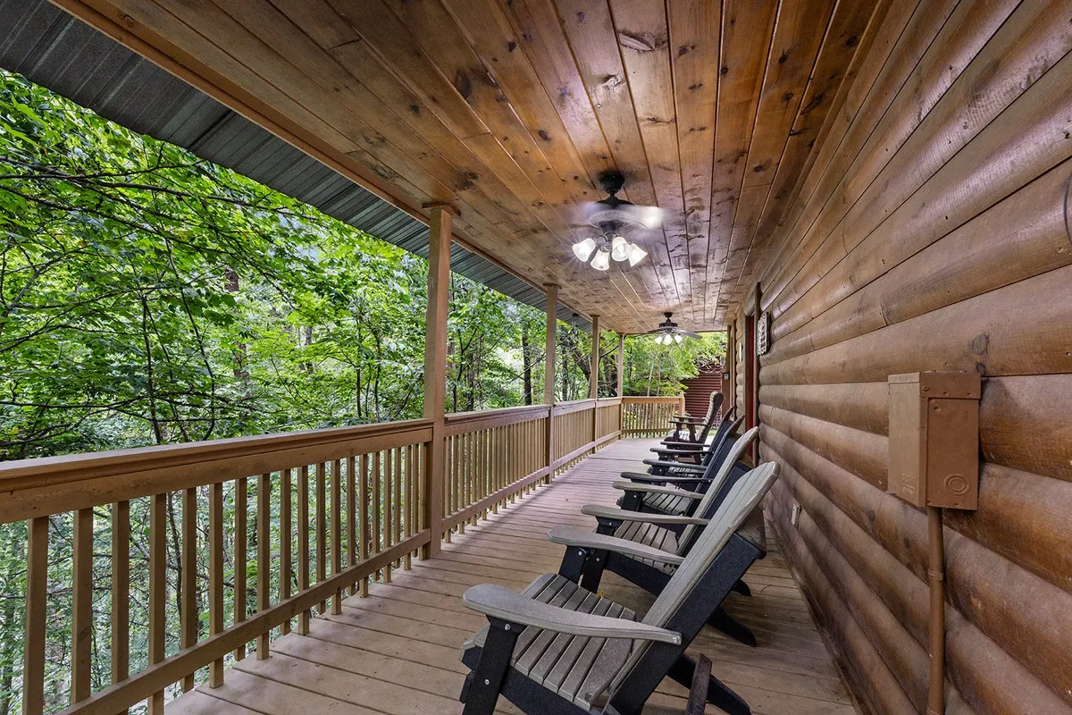 A covered porch with sturdy wooden railings, several classic rocking chairs, a ceiling fan overhead, and lush greenery all around. The porch is bathed in soft natural light, inviting guests to sit back and soak in the tranquil mountain setting and forested views.