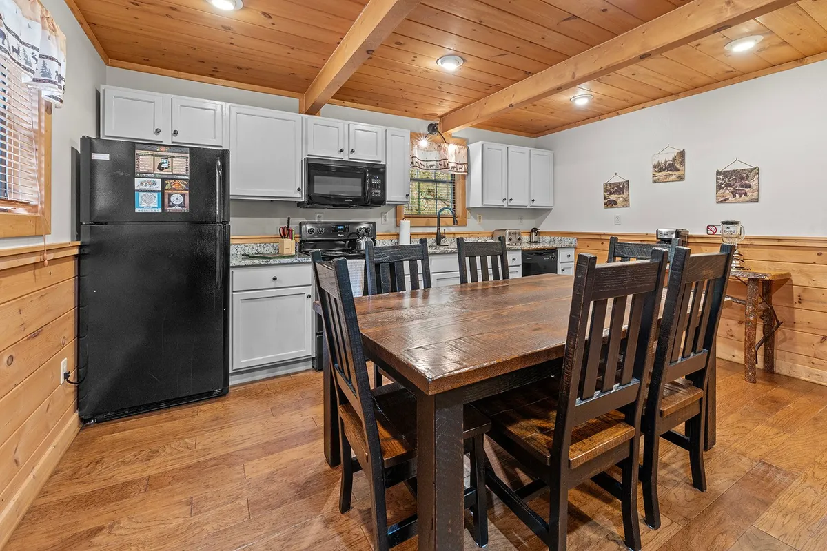 A close look at the spacious dining table and adjacent kitchen in a log cabin setting. Wooden beams and cabinetry frame the room, with modern appliances and a well-lit dining space ready for shared meals or board games. The layout emphasizes communal dining and kitchen activity.