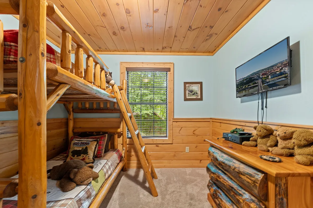 A cheerful, kid-friendly room featuring a wooden bunk bed, TV mounted on the wall, a wooden dresser, and stuffed bears. The space feels playful yet cozy, designed for both rest and entertainment, appealing especially to families with children.