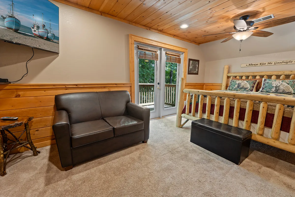 A spacious bedroom with a log king bed, sliding glass doors leading to a private balcony, and a leather loveseat. Soft natural light fills the room, and nature views can be seen through the glass doors, creating a tranquil, restful setting. Artwork on the wall and comfortable furnishings add to the suite-like feel.