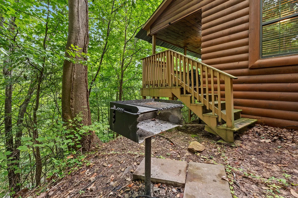 A cozy outdoor grill area featuring a charcoal grill set on a wooden deck, bordered by wooden steps and natural greenery. The cabin’s exterior is visible, blending seamlessly into the forested landscape, creating an inviting spot for social barbecues and outdoor meals.