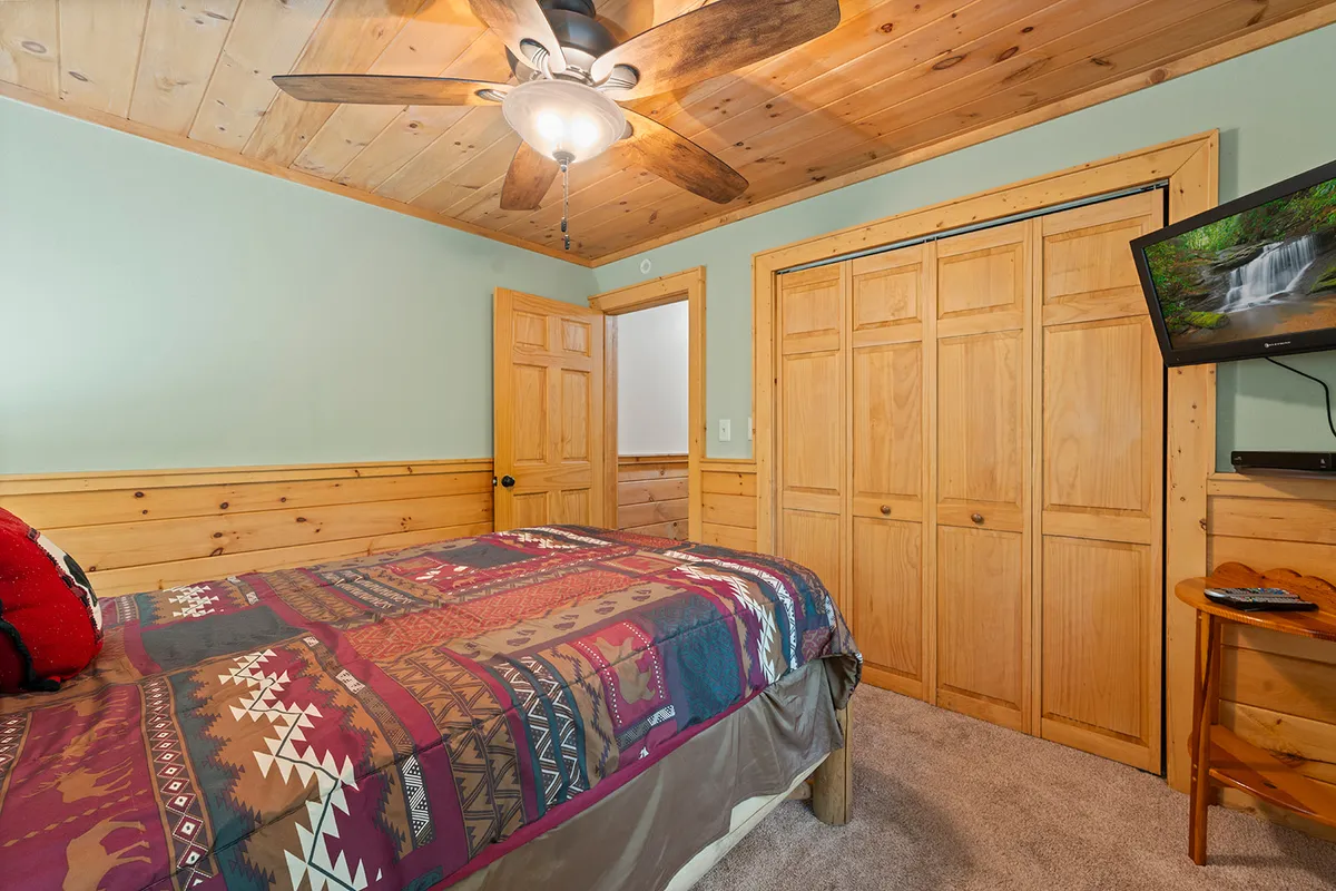 A comfortable bedroom with a bed dressed in a patterned blanket, a wooden ceiling fan, light-colored walls, wooden closet doors, and a TV mounted on the wall. The room exudes a rustic yet inviting atmosphere, perfect for resting after a day of adventure.