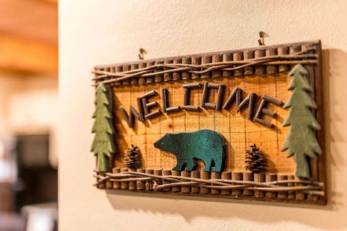 A rustic wooden welcome sign featuring a bear silhouette and pine trees, positioned near the cabin entrance. The natural textures and playful design evoke both the mountain setting and the spirit of exploration.