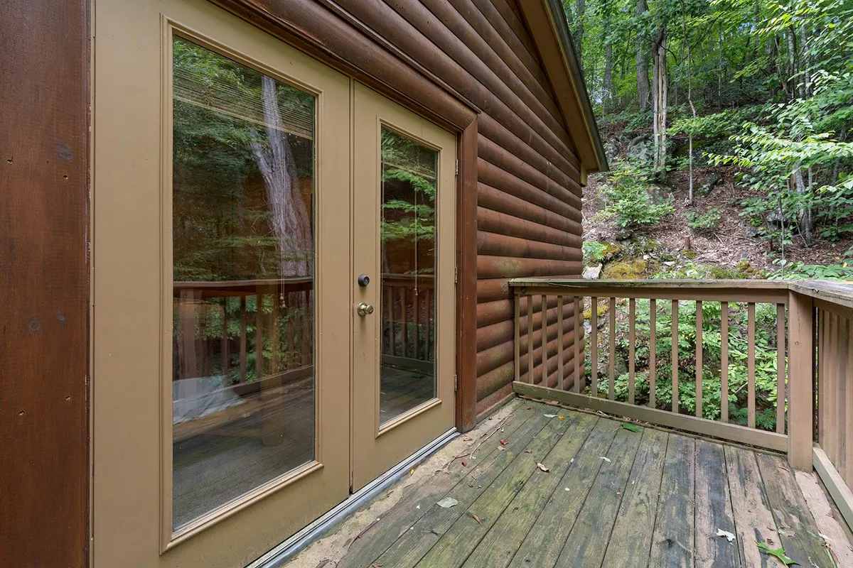 This image showcases the exterior wooden deck of the cabin, with double glass doors opening to lush greenery. The scene is set for relaxation, with ample space for enjoying nature, sipping morning coffee, or stargazing at night.