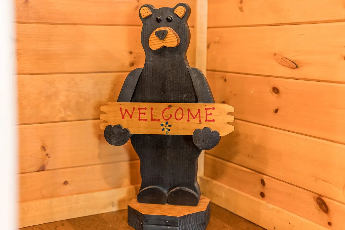 A close-up of a charming wooden bear statue holding a 'WELCOME' sign, set against the background of a warmly lit entryway. The carved bear adds a playful, inviting touch, signaling a friendly arrival and the start of a memorable getaway.