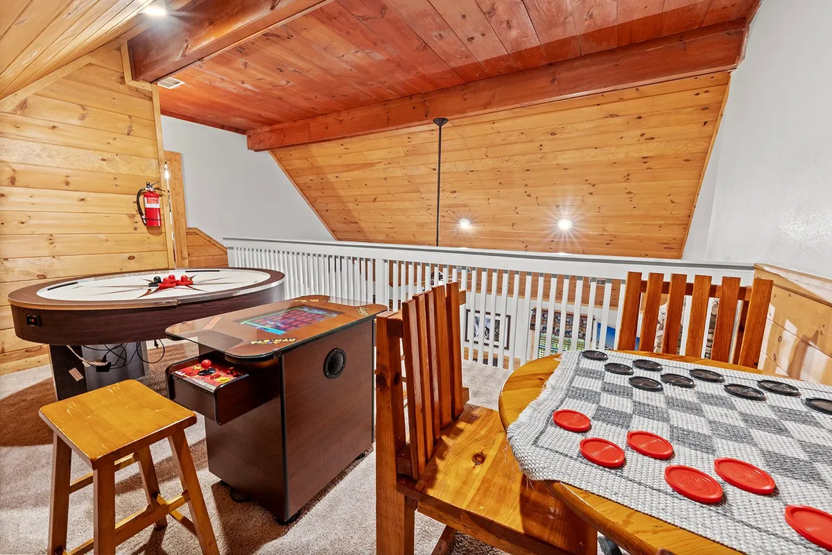 This image features a game table set up for play, an arcade machine in the background, a checkers board, and wooden chairs arranged for group fun. The space is designed for interactive entertainment and socializing, with a cozy cabin feel.