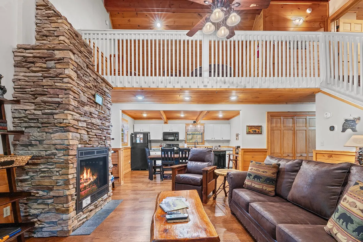 A warm, open-concept living and dining area featuring a rustic stone fireplace, wooden ceilings, a plush sofa, and a wooden dining table set for a group. The kitchen area is visible in the background, seamlessly blending with the dining and lounging spaces. Sunlight filters through large windows, creating an inviting atmosphere ideal for gatherings.