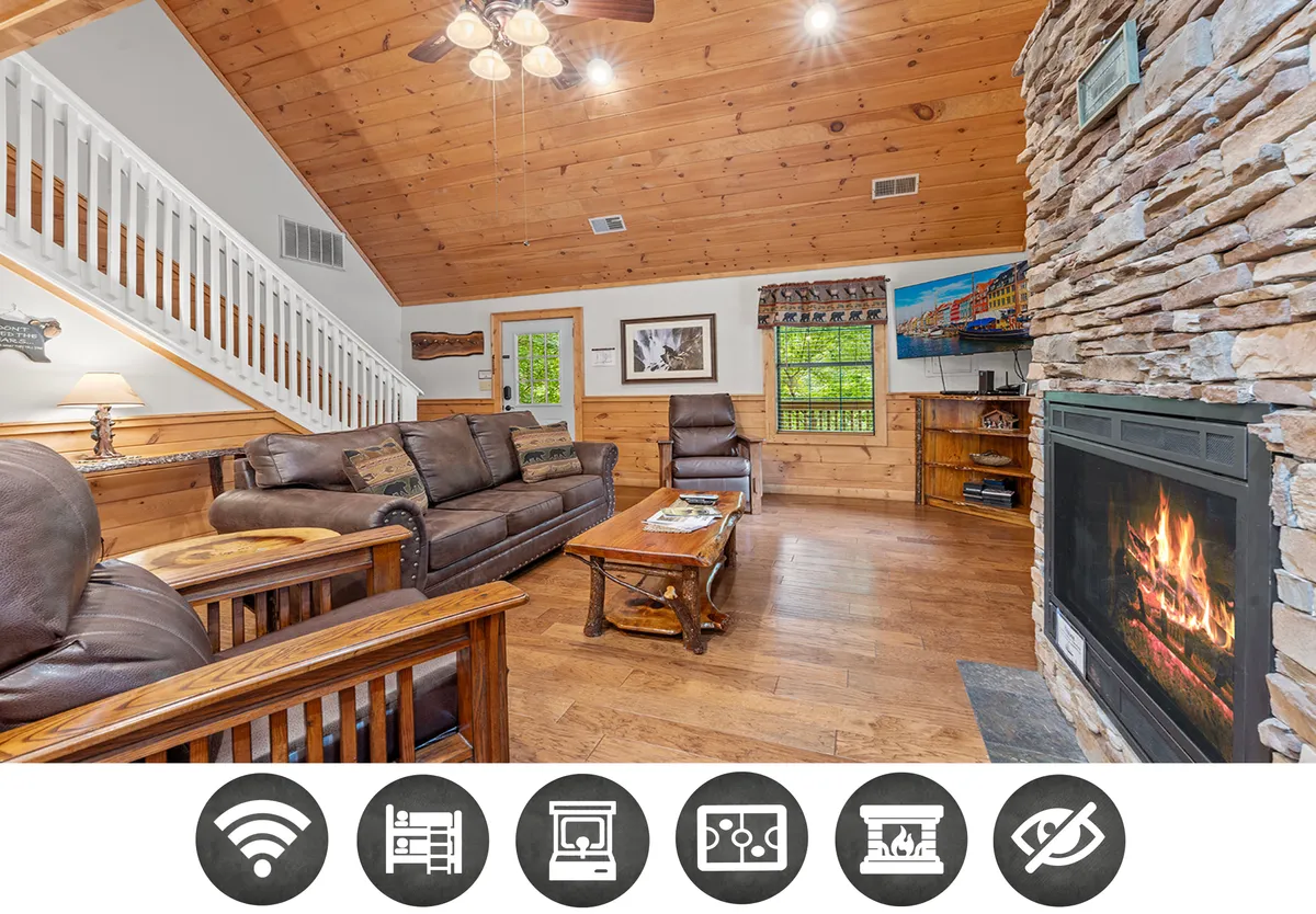 The image showcases a warm and inviting living area featuring a stunning stone fireplace as the focal point, surrounded by rustic wooden furniture. Large windows bring in natural light and offer glimpses of the tranquil mountain scenery. The ambiance is cozy, with a ceiling fan overhead and comfortable seating ideal for family game nights, quiet reading, or relaxing by the fire.