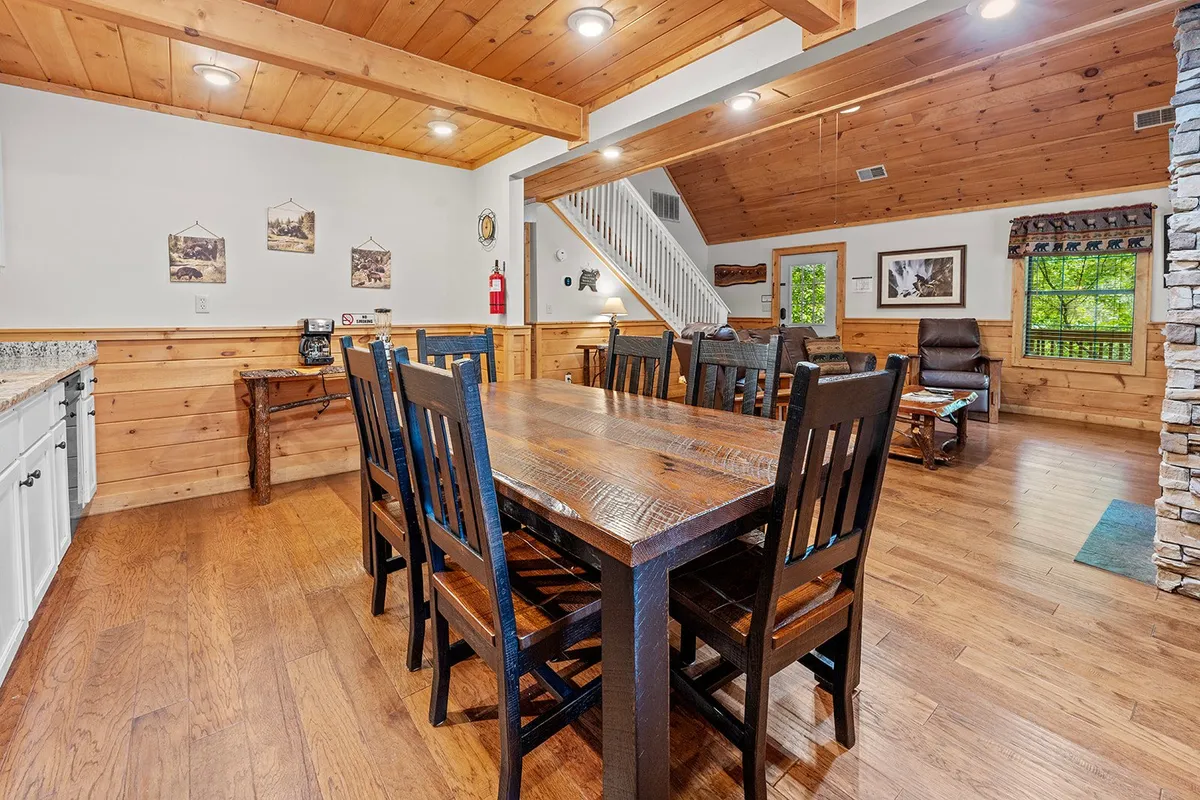 A cozy interior with a large wooden dining table, wooden chairs, a stone fireplace, and a staircase leading to the upper level. The space is arranged for group dining, game nights, and casual gatherings, radiating warmth and togetherness.
